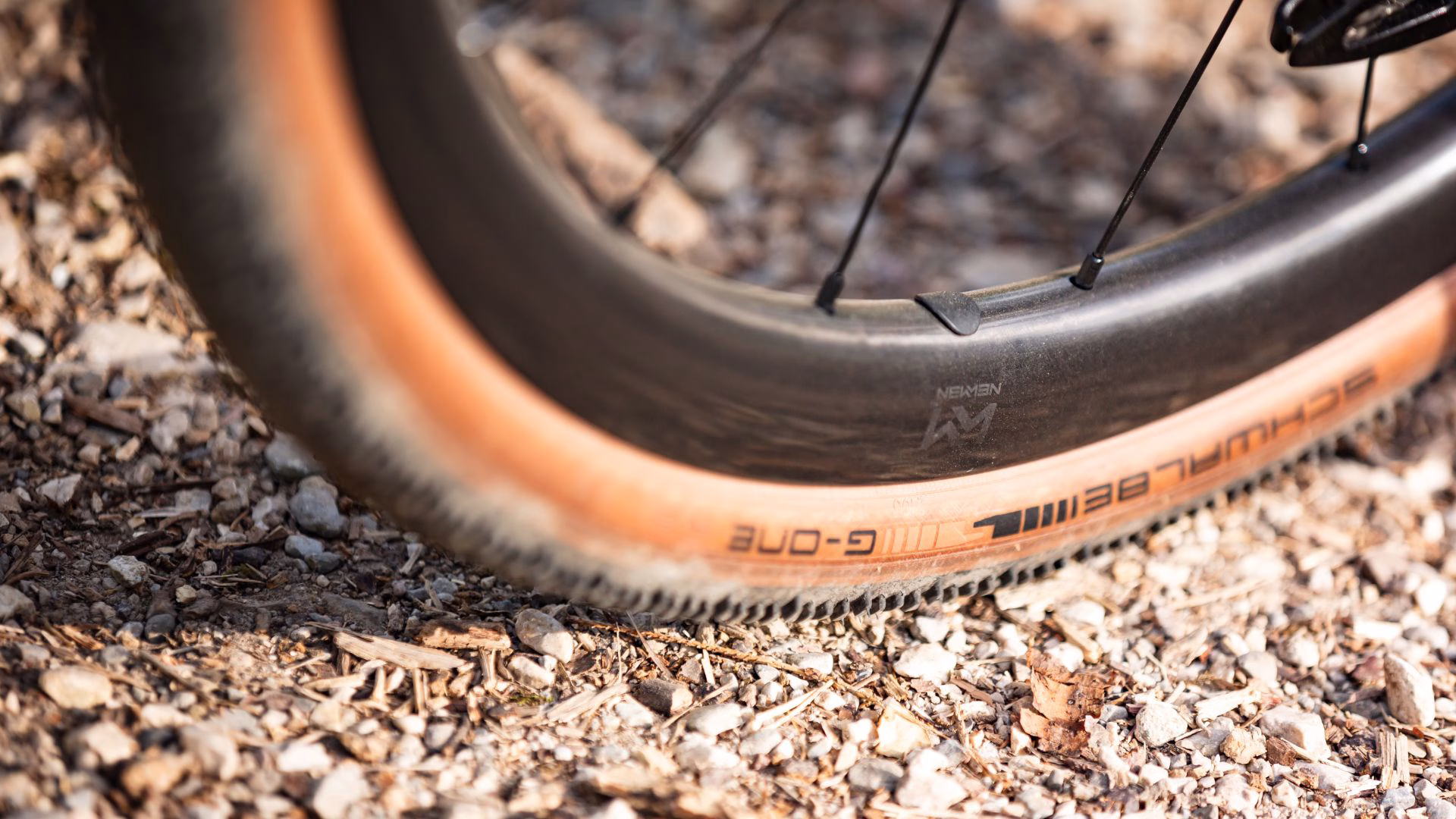 Close-up of a Schwalbe G-One gravel tire with tan sidewall on a carbon Newmen rim above a rocky gravel path.