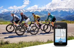 Vier racefietsers rijden over een bergweg met besneeuwde toppen, met daarnaast een Garmin Edge 1060 die een melding over een gat in de weg toont