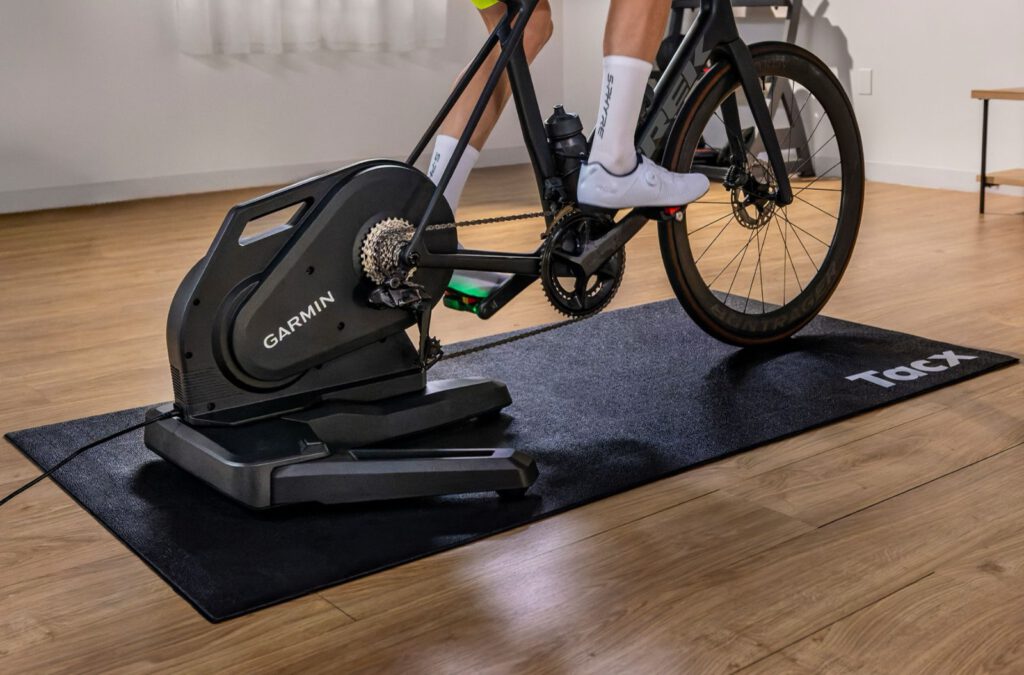 Racefiets op de Garmin Tacx Nēra smart trainer op een zwarte Tacx-mat in een woonkamer met houten vloer