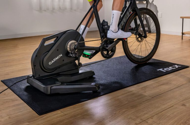 Racefiets op de Garmin Tacx Nēra smart trainer op een zwarte Tacx-mat in een woonkamer met houten vloer