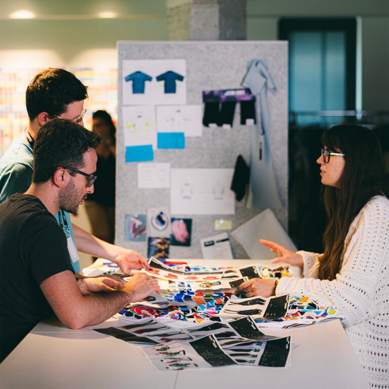 Ontwerpers van Santini bespreken kleurstalen en shirtontwerpen aan een tafel vol prints