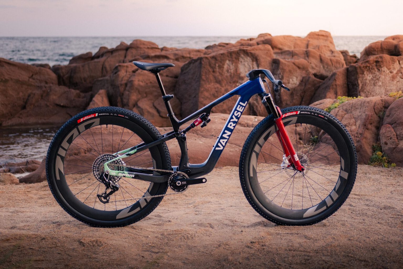 De Van Rysel RCR-XC mountainbike in blauw en zwart met rode vork, op een rotsachtige kust bij zonsondergang