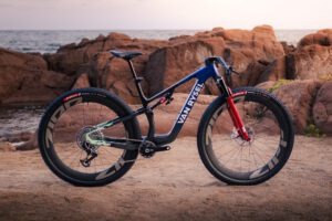 De Van Rysel RCR-XC mountainbike in blauw en zwart met rode vork, op een rotsachtige kust bij zonsondergang